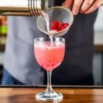 A bartender pouring a pink Shaken strawberry daiquiri mocktail through a fine mesh strainer into a glass